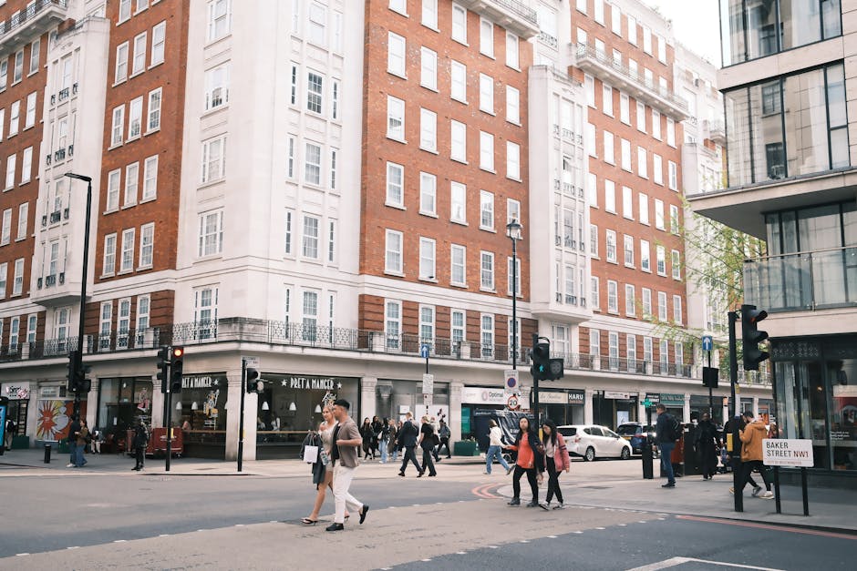 Urban street scene showing a multi-story brick and white plaster building with large windows, housing retail stores at ground level such as Pret A Manger and other shops. The sidewalk is bustling with pedestrians crossing the road, some carrying shopping bags or using smartphones. Several traffic lights and street lamps are visible, along with a street sign indicating Baker Street NW1. On the right, a modern building with glass balconies reflects daylight, enhancing the city atmosphere. The scene captures the lively atmosphere of Marylebone, highlighting the importance of surface cleaning and maintenance in busy commercial areas, which is a service provided by Cleaners Marylebone for deep cleaning and sanitisation of residential and commercial spaces.