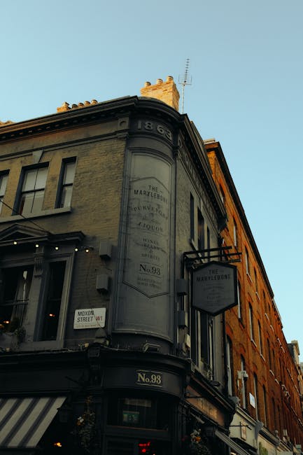 Photograph of a historic building with a rounded corner facade, featuring a large vertical sign reading 'The Marylebone'. The building’s exterior is composed of light-colored stone or brick, with decorative architectural details along the roofline and cornices. The ground floor has large windows, possibly storefronts, with striped awnings. The image captures clear daylight with a pale blue sky, and the scene reflects a clean and well-maintained appearance, aligning with professional domestic and commercial surface cleaning standards promoted by Cleaners Marylebone. Visible signage indicates the building’s address as No. 98, located in Marylebone W1U, an area known for its historic architecture and retail establishments.